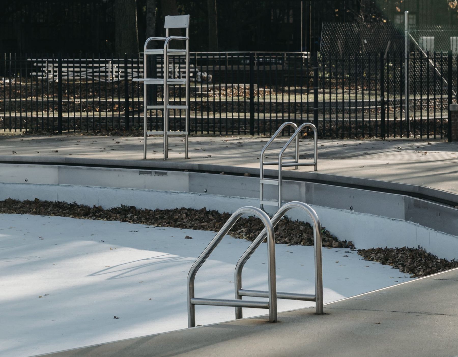 A drained municipal swimming pool out of season, with an empty lifeguard chair, stainless handrails and fallen leaves collected in the gutter.